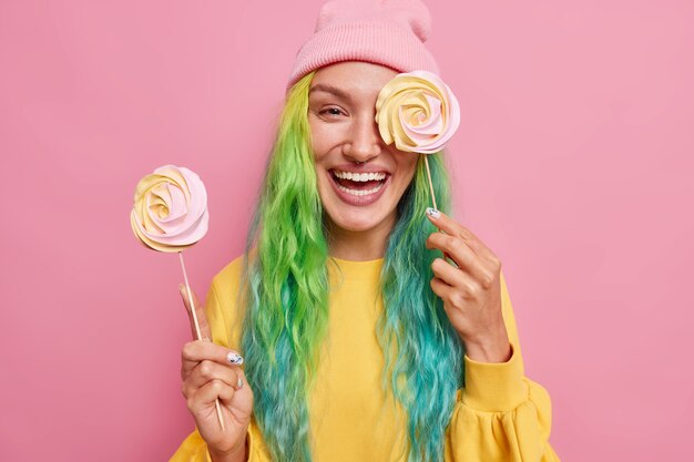  woman holds two round candies on sticks cons eye with delicious caramel lollipop has colorful hair wears yellow jumper and hat isolated on pink 