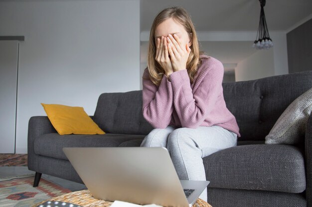 Woman at home closing eyes with hands, thinking. Laptop on table