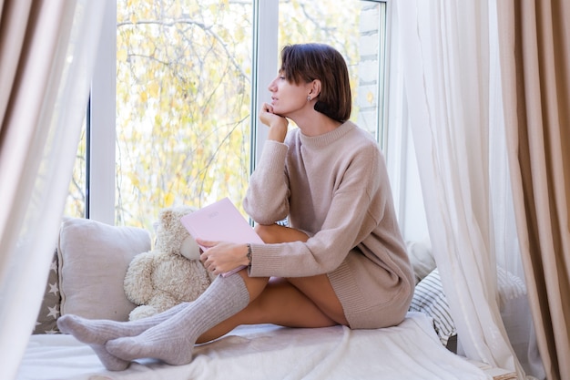 Woman at home with notebook sit on windowsill in comfy sweater and warm wool socks, cold outumn outside window
