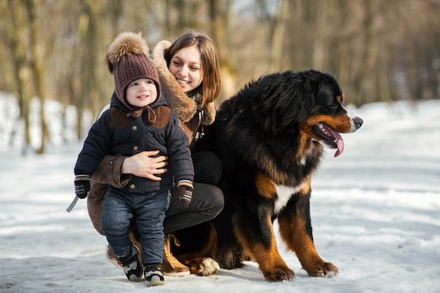 Woman hugs little boy and strokes Bernese Mountain dog posing in the park