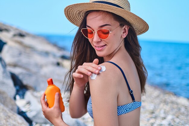 水着 麦わら帽子 真っ赤なサングラスを着て幸せな女は 夏の晴天の海でリラックスして海で日光浴をしているときに彼女の肩に日焼け止めを塗る プレミアム写真