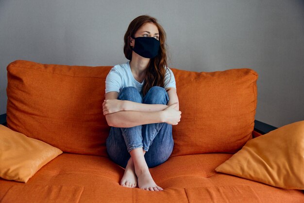 黒​の​医療​マスク​の​女性​が​ソファ​で​自宅​で​ロック​ダウン​滞在​コンセプト