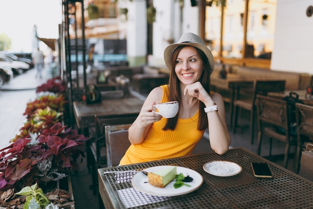 카푸치노, 케이크 한잔과 함께 노란색 옷 모자에 테이블에 앉아 야외 거리 커피 숍 카페에서 여자, 자유 시간 동안 레스토랑에서 휴식
