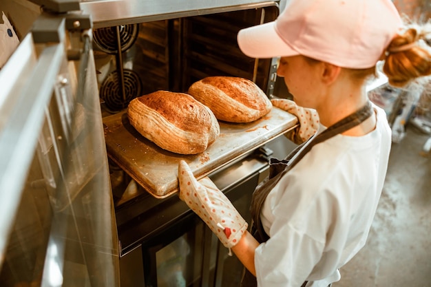분홍색 모자를 쓴 여성이 공예 베이커리 작업장에서 오븐에서 빵을 꺼냅니다.