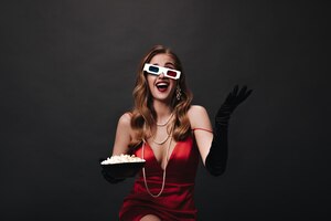 Женщина в красном платье в 3d-очках и с попкорном в руках леди в шелковом праздничном наряде смотрит фильм на черном фоне