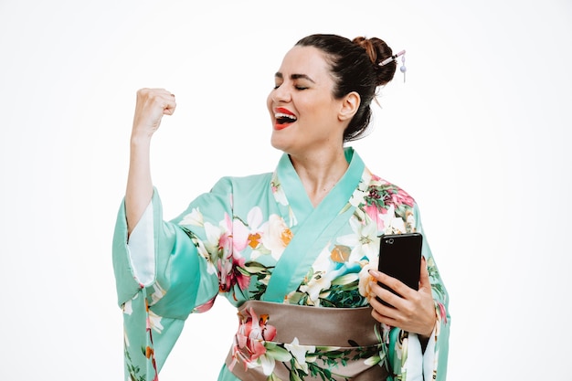 白で幸せで興奮して彼女の成功を喜んで拳を握りしめるスマートフォンを保持している伝統的な日本の着物の女性