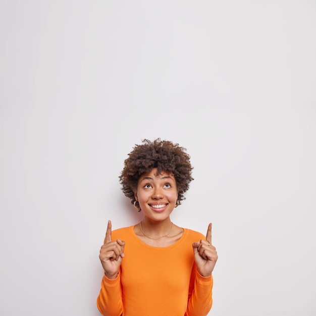 женщина показывает выше двумя указательными пальцами, носит повседневный оранжевый джемпер, привлекает ваше внимание крутым предложением, изолированным на белом фоне, показывает логотип