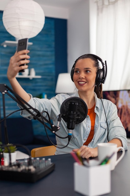 Woman influencer in home studio streaming live on smartphone. Content creator using microphone and mobile phone to stream and record for social media channel. Podcast professional home studio.