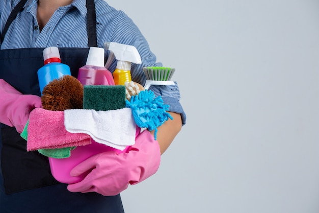 Woman is holding cleaning product, gloves and rags in the basin on white wall