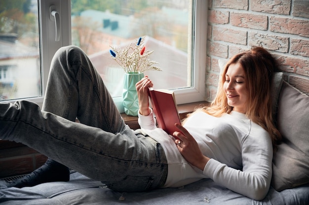 Woman is lying on windowsill bed and reading book