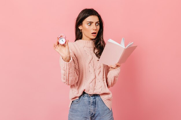 Woman is reading shocking book and holding pink alarm clock. Shot of lady in sweater and jeans on isolated background.