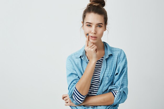 Woman is trying to think up where to put new sofa. Gorgeous modern woman with bun hairstyle holding hand on chin while looking with focused and determined expression  