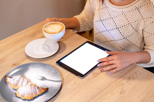 A woman is using a tablet holding a cup of coffee, dessert on a wooden table