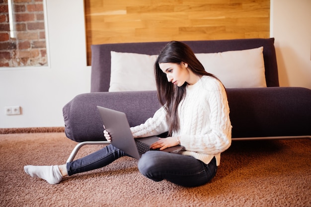 Free photo woman is working on laptop at home sitting on the floor near the sofa in white sweater