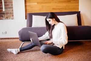 Бесплатное фото Женщина работает на ноутбуке дома сидит на полу возле дивана в белом свитере