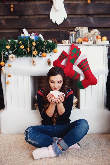 クリスマスのもので飾られた暖炉の前に、ジーンズの女性がホットドリンクのカップと座っている
