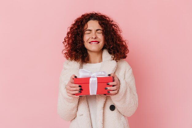 Woman in joyful anticipation holds gift. lady with dark curly hair on pink space.