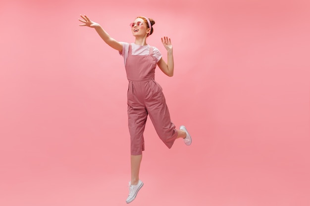 woman jumping on pink background. Full-length shot of redhead woman in bright outfit and glasses.