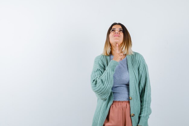 Woman keeping finger under chin in casual clothes and looking jolly , front view.