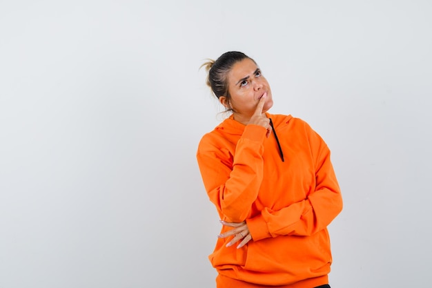 woman keeping finger on lips in orange hoodie and looking pensive