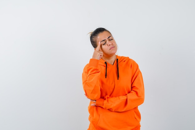 Woman keeping finger on temples in orange hoodie and looking pensive