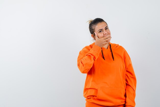 woman keeping hand on mouth in orange hoodie and looking cheery