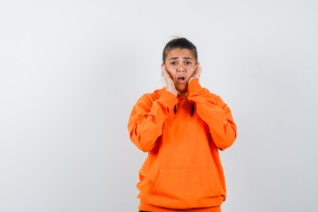 woman keeping hands on cheeks in orange hoodie and looking anxious