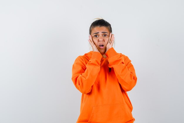 woman keeping hands on cheeks in orange hoodie and looking anxious