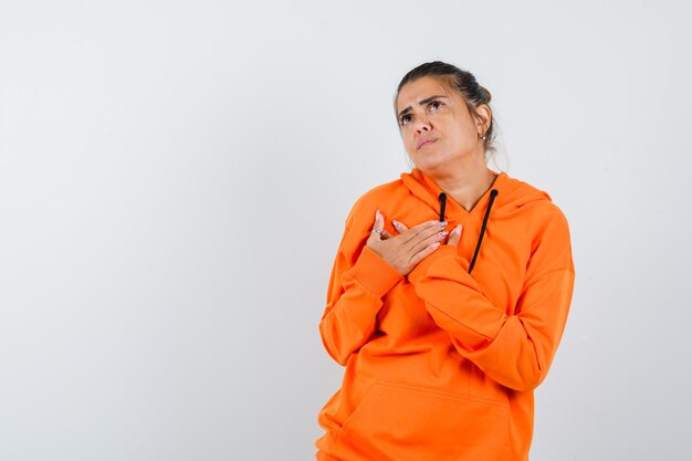 Женщина, держащая руки на груди в оранжевой толстовке с капюшоном и задумчивая