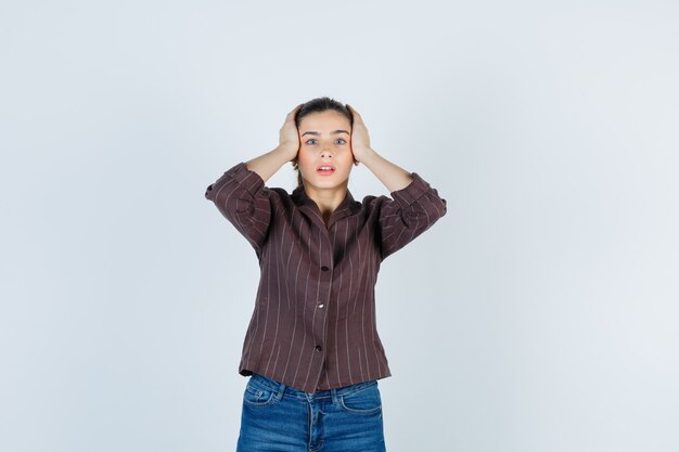 woman keeping hands on head in shirt, jeans and looking agitated , front view.