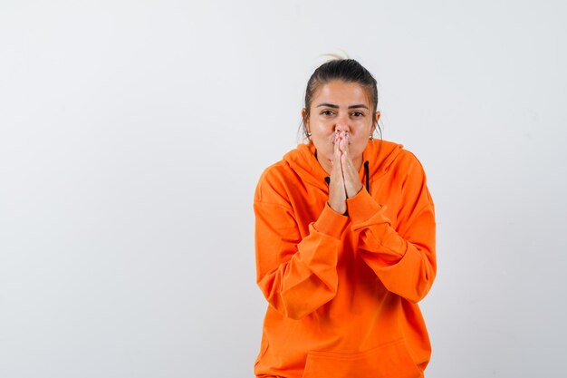 Woman keeping hands in praying gesture in orange hoodie and looking hopeful