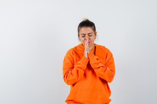 Woman keeping hands in praying gesture in orange hoodie and looking hopeful