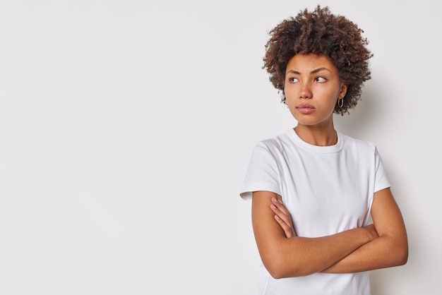 woman keeps arms folded concentrated away with serious expression dressed in casual t shirt isolated on white copy space area for advert