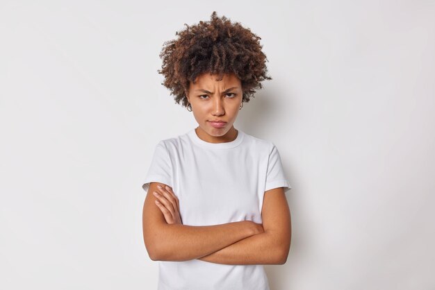 woman keeps arms folded feels offended or insulted frowns from under forehead looks with disbelief and offence wears casual t shirt isolated on white.