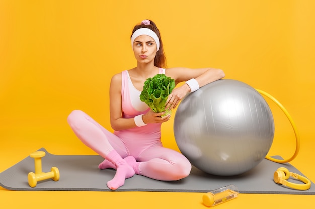 女性はダイエットを続けます健康を保つための定期的なフィットネストレーニングを持っていますスポーツ服に身を包んだ緑の野菜を保持しますヘッドフォンでマットの上に座っていますダンベルスイスボールフラフープジムで