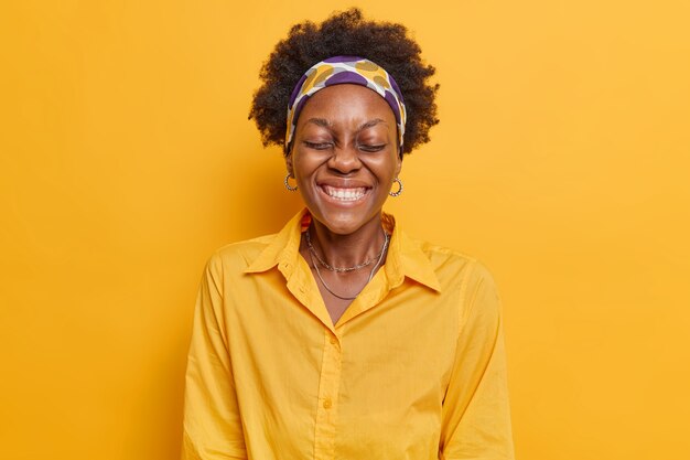 Woman keeps eyes closed laughs happily chuckles carefree shows white teeth wears headband and shirt isolated on vivid yellow