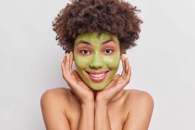女性は顔に手を保ち、白地にトップレスの肌に栄養を与えるポーズに緑のキュウリマスクを適用します