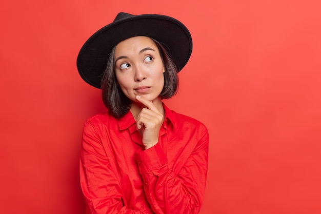 Foto gratuita la donna tiene il mento di handon immerso nei pensieri considera che qualcosa indossa una maglietta nera alla moda che posa sul rosso