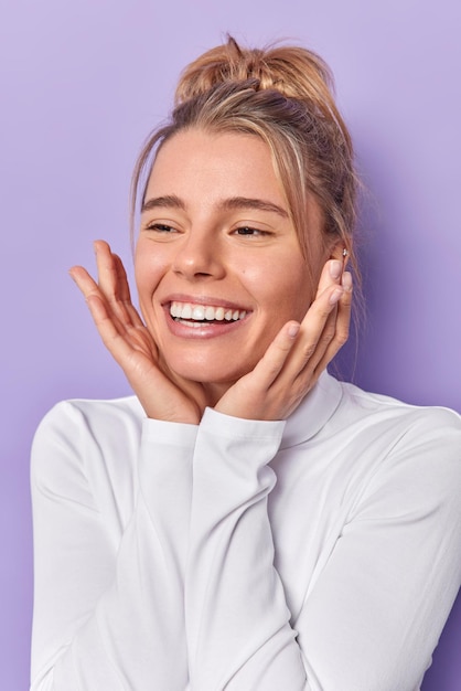 Free photo woman keeps hands on cheeks smiles broadly with white teeth dressed casually isolated on purple. people happy emotions and face expressions concept
