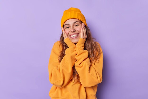 woman keeps hands on cheeks smiles toothily has glad expression dressed in orange casual jumper and hat isolated on purple. Pretty millennial girl feels very happy