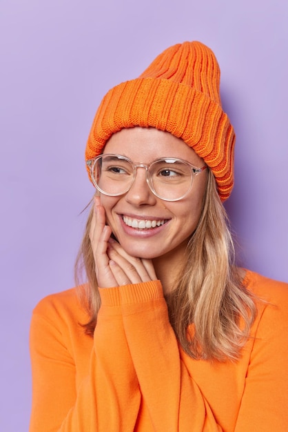女性は顔の近くで手を優しく保ち、歯は目をそらし、オレンジ色の長袖のジャンパーを着ており、紫色で隔離された帽子は楽しい考えを持っています。幸せな感情
