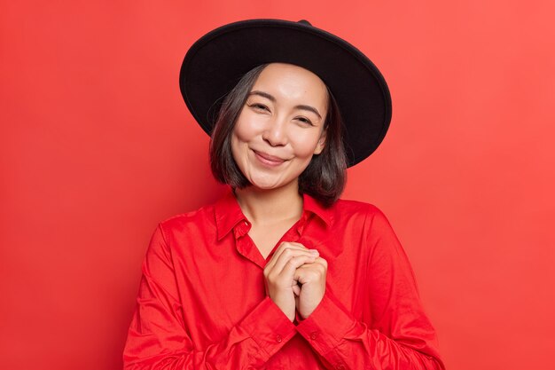  woman keeps hands pressed together near heart feels touched and pleased smiles gently at camera wears black hat shirt stand on vivid red 