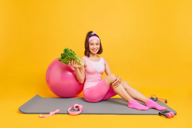 woman keeps to healthy diet holds green fresh vegetable sits on mat poses around sport equipment 