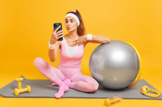 woman keeps lips folded makes selfie on smartphone or has video conversation dressed in sportswear poses sits at fitness mat surrounded by sport equipment isoated on yellow wall