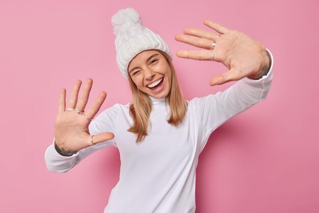 woman keeps palms forward camera winks eyes and smiles gladfully wears white winter hat and turtleneck looks happily at camera isolated on pink. Positive emotions