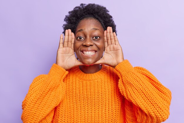  woman keeps palms near face smiles broadly eyes full of happiness foolishes around wears orange knitted jumper isolated on purple 