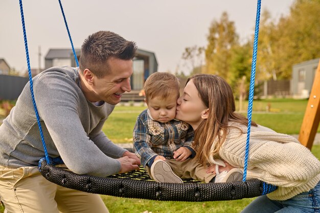 Woman kissing child sitting on swing and man watching