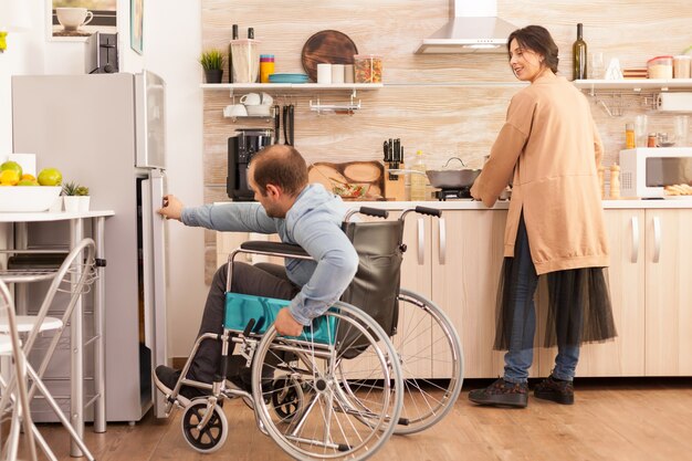 Woman in kitchen looking at husband with walking disability trying to open refrigerator door. Disabled paralyzed handicapped man with walking disability integrating after an accident.