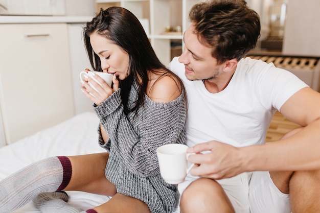 彼女のボーイフレンドの横にコーヒーのカップと一緒にベッドに座っているニットの灰色のセーターの女性