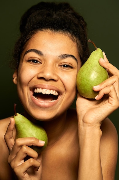 woman laughing at camera, holding two juicy pears near her face, posing isolated on green